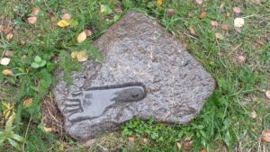 Taideteoksen osa, jossa kiveen näyttää painuneen ihmisen jalanjälki. Kuvattuna kesällä, kiven ympärillä on vihreää ruohoa.